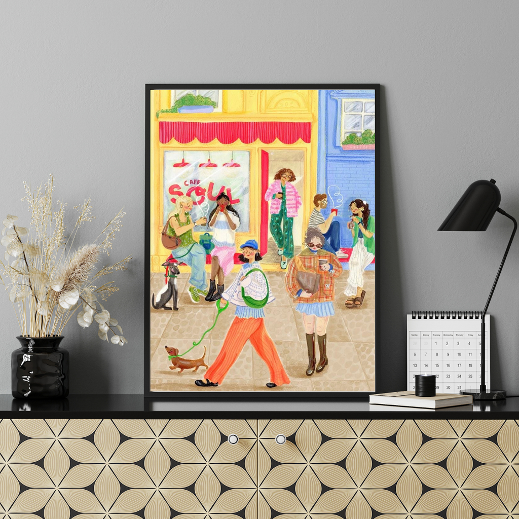 Framed artwork of people on a street with a store front, displayed on a wall above a decorative shelf.